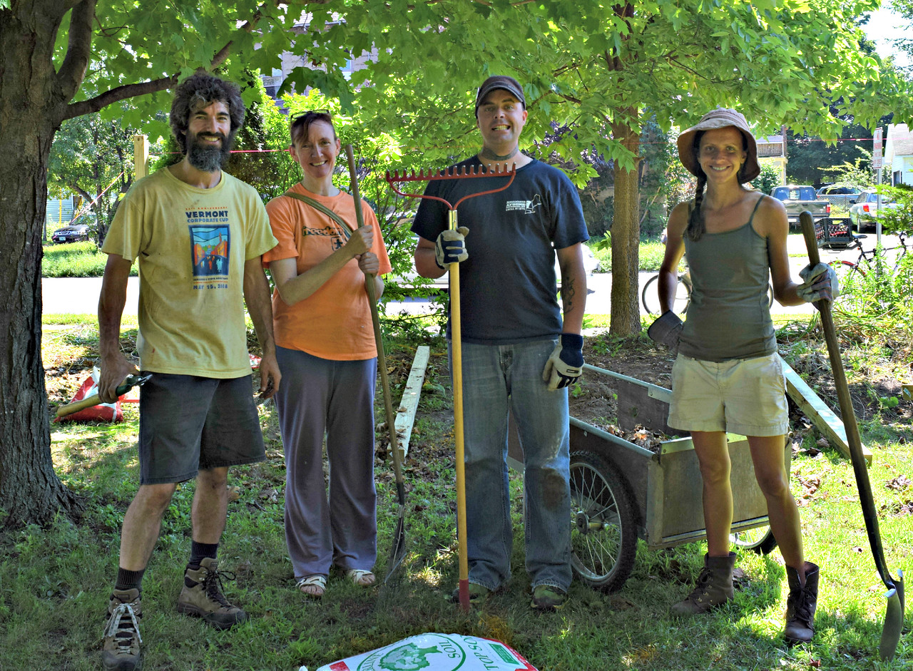 Four people are standing outside together holding gardening tools