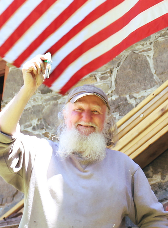 A man smiling holding a key. He is wearing a gray sweatshirt and hat and standing in front of an American flag.