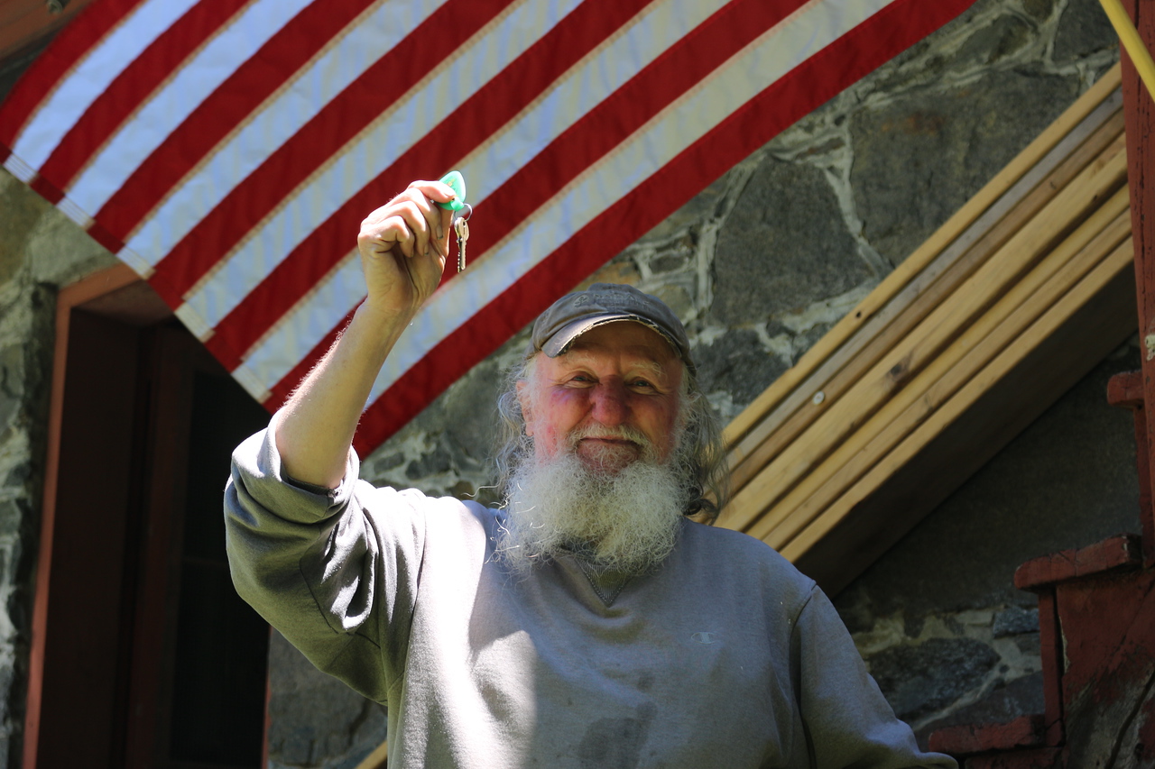 A man smiling holding a key. He is wearing a gray sweatshirt and hat and standing in front of an American flag.