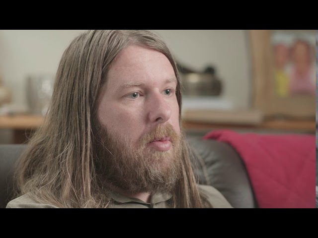 The person in the photo has long, dirty blonde hair. They aren't looking straight at the camera. They have a beard and are sitting on a brown couch with a red blanket in the background.