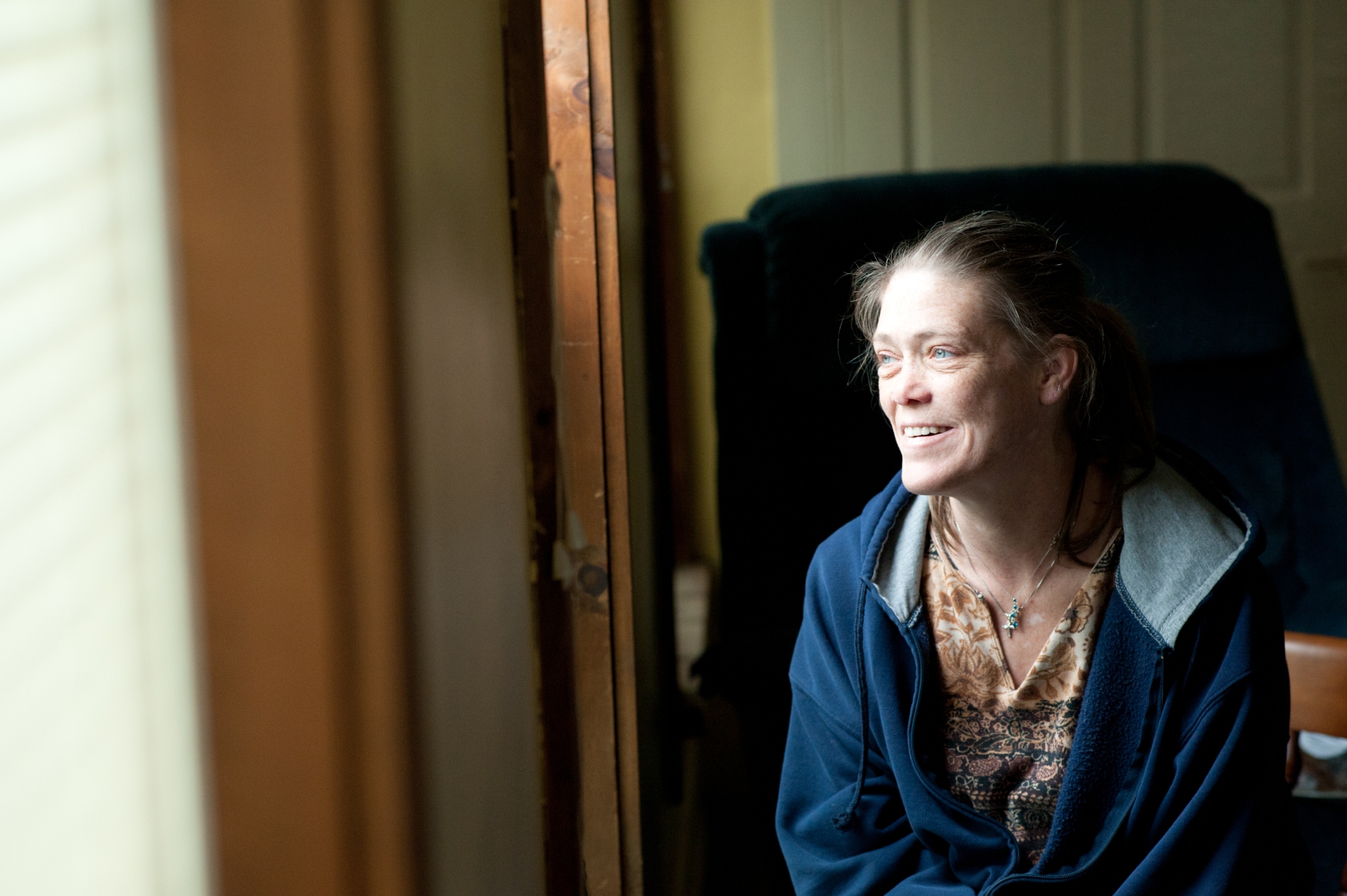 A woman looking out a window. She is smiling and wearing a blue sweater with a brown patterned shirt underneath. She has blue eyes.