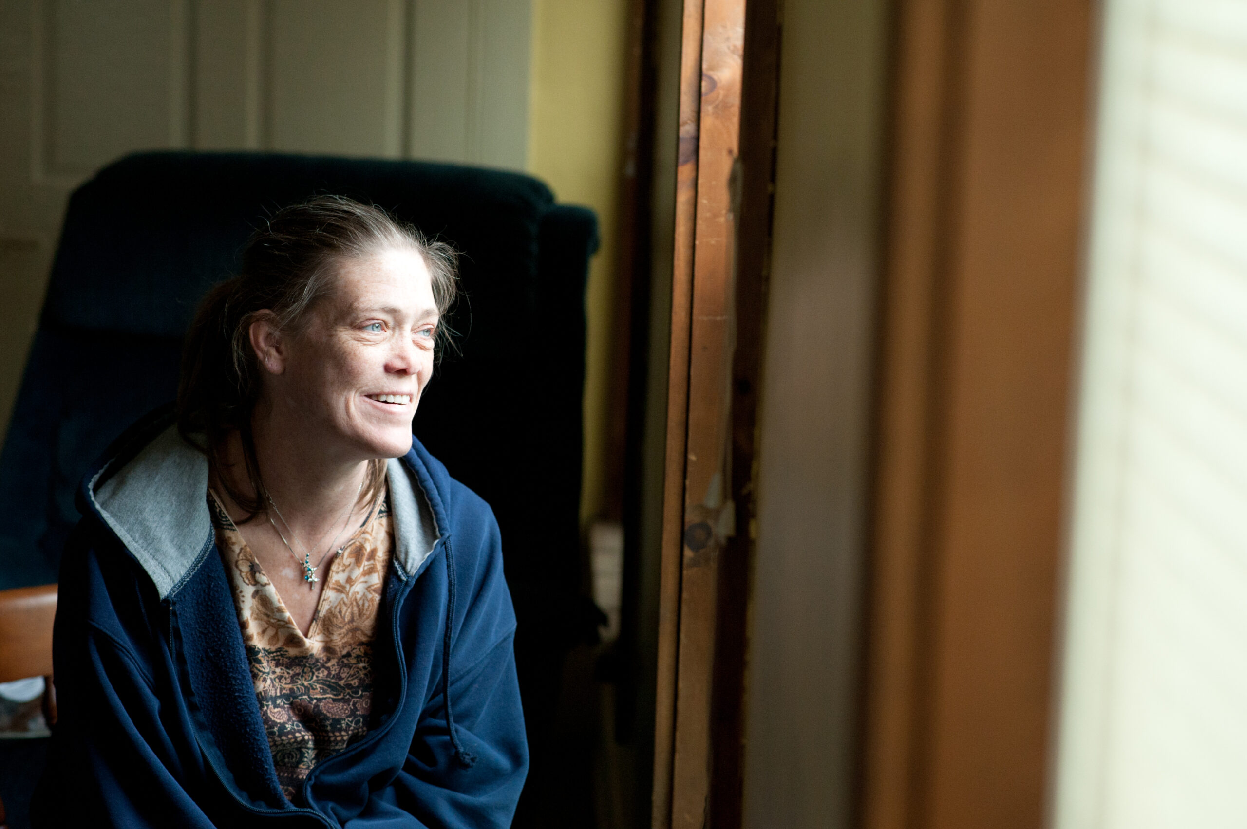 A woman looking out a window. She is smiling and wearing a blue sweater with a brown patterned shirt underneath. She has blue eyes.