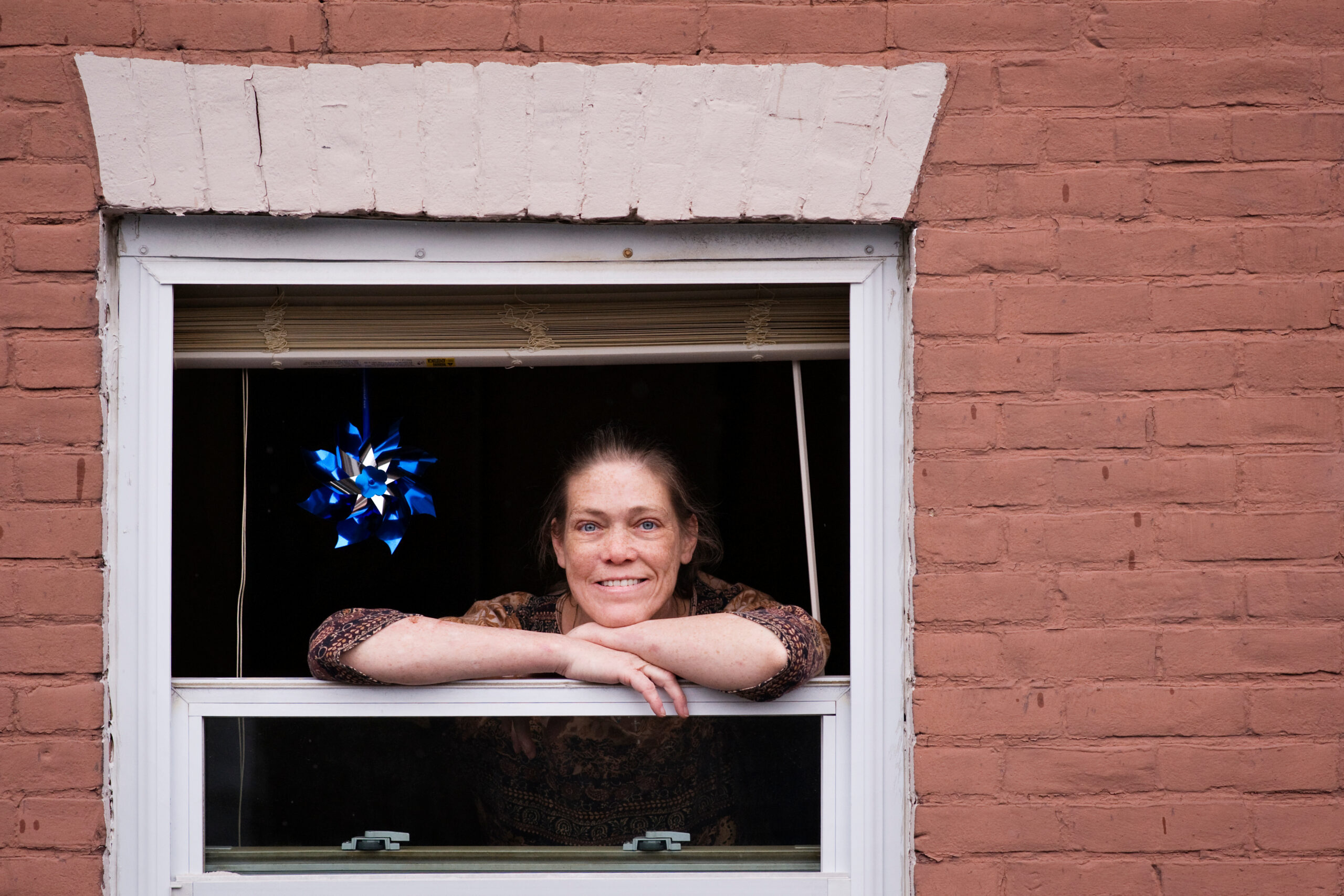 A woman looking out of a window. There is a blue ribbon hanging to her left and she is smiling.