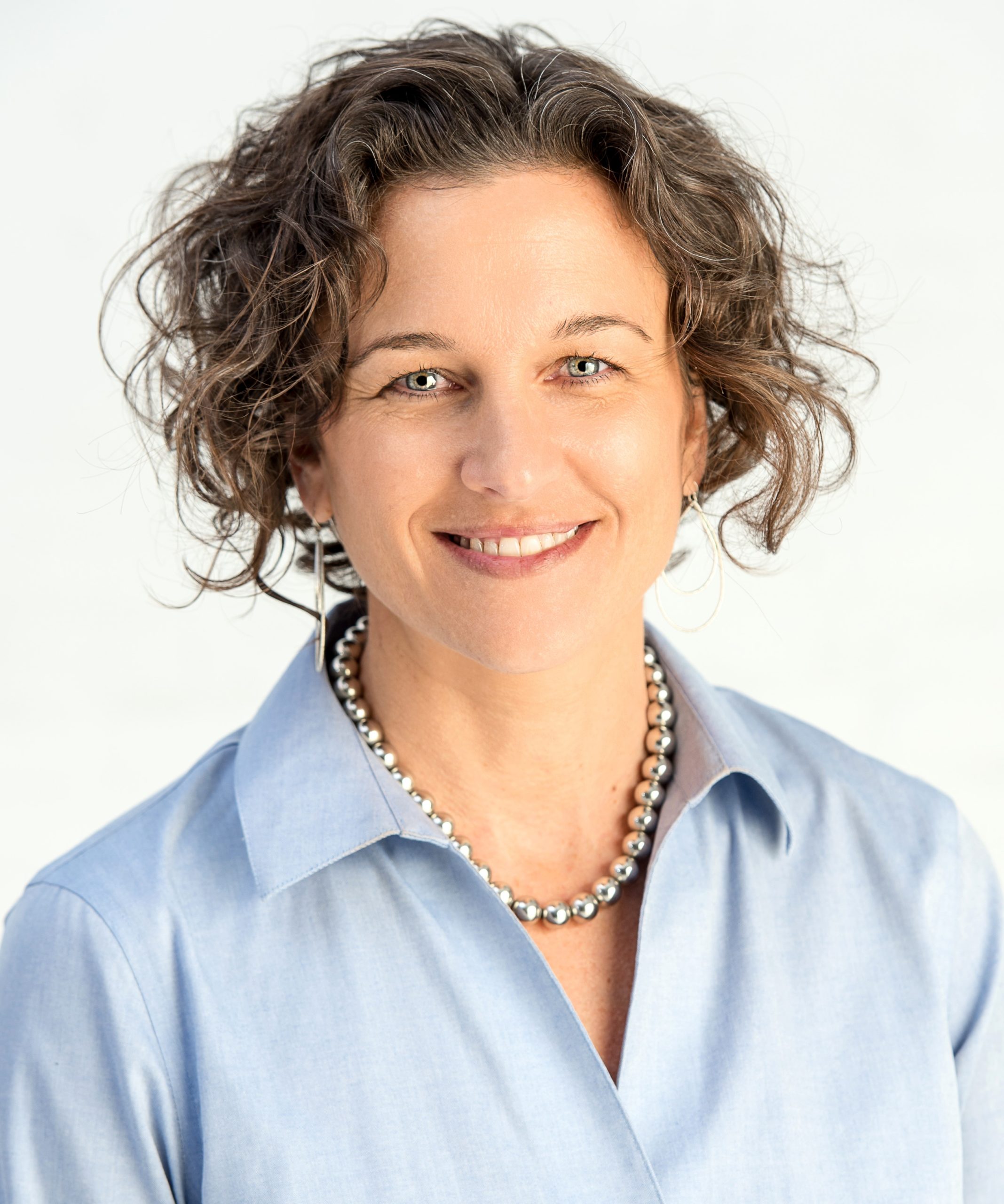 Photo of a woman wearing a blue blazer, a large silver necklace, and earrings. She has short, curly brown hair and is smiling.