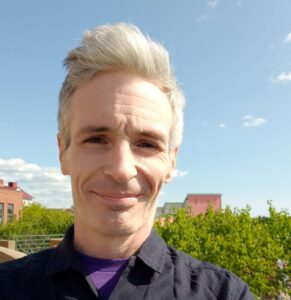 Selfie of a man wearing a purple shirt and black jacket. There are trees and the tops of buildings in the background.