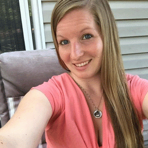 Selfie taken of a woman wearing a pink shirt and necklace. She has long, light brown hair and is sitting on a chair on a porch.