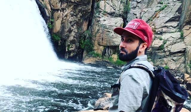 Photo of a man with a beard standing by a waterfall wearing a denim shirt, red shorts, a backpack, and a red hat.