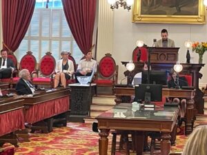Photo of a man presenting at a podium at the Vermont statehouse. There are six other people in the photo who are looking at the presenter. The interior is red and brown.