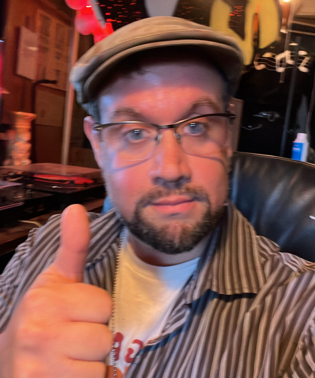 Selfie of a man wearing glasses who is doing a thumbs up for the camera. He is wearing a hat, a striped brown overshirt, and a white undershirt. He is sitting on a leather chair with a record player in the background.