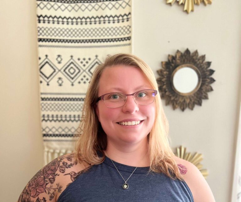 Photo of a woman wearing a blue tank top, a necklace, glasses and a nose stud. She has tattoos on her left shoulder.