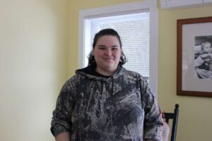 Photo of a woman wearing a camouflage sweatshirt with brown hair and a lip ring. She is standing in a room with beige walls and a photograph of a baby