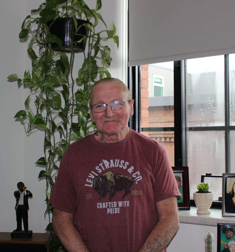 Photo of a man with glasses wearing a red "Levi Strauss & Co" shirt that has a bison and says "Crafted with Pride." He is standing in front of plants and photos of his family.