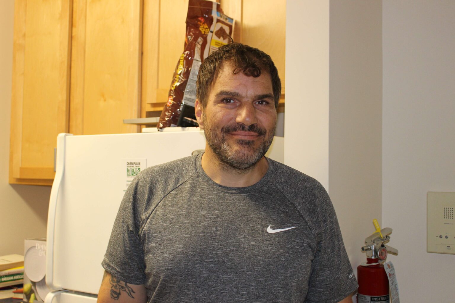 Photo of a man wearing a gray Nike t-shirt with short brown hair. He has a tattoo on his left arm and is standing in the entrance to his kitchen
