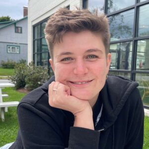 Photo of a woman smiling wearing a black sweatshirt. She is wearing earrings and a ring. In the background is a white picnic table and a building with glass windows.
