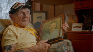 Photo of a man wearing a yellow shirt and a black hat that says "World War II Veteran." He is holding a photo of a woman wearing a blue shirt. The background is his home, with other photos and decorative pieces.