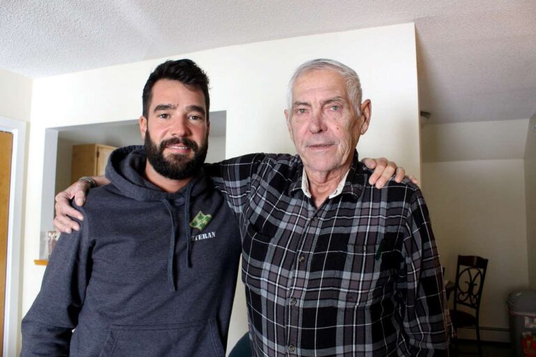 Photo of two men standing smiling at the camera. The man on the left is wearing a navy blue sweatshirt that says "Veteran" and the man on the left is wearing a black and gray flannel.