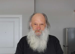 Photo of a man with a long beard who is wearing a black long sleeve top. He is standing with his kitchen in the background.