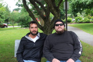 Photo of two men sitting on a park bench. The man on the left has a beard and is wearing a white shirt and a black raincoat. The man on the right also has a beard and is wearing a black long sleeve shirt, sunglasses, and a black beanie. In the background are trees and a path through the park.