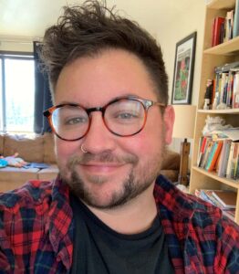 Selfie of an individual wearing a black shirt and red flannel. They are also wearing glasses and have a nose ring. In the background is a bookshelf and couch.