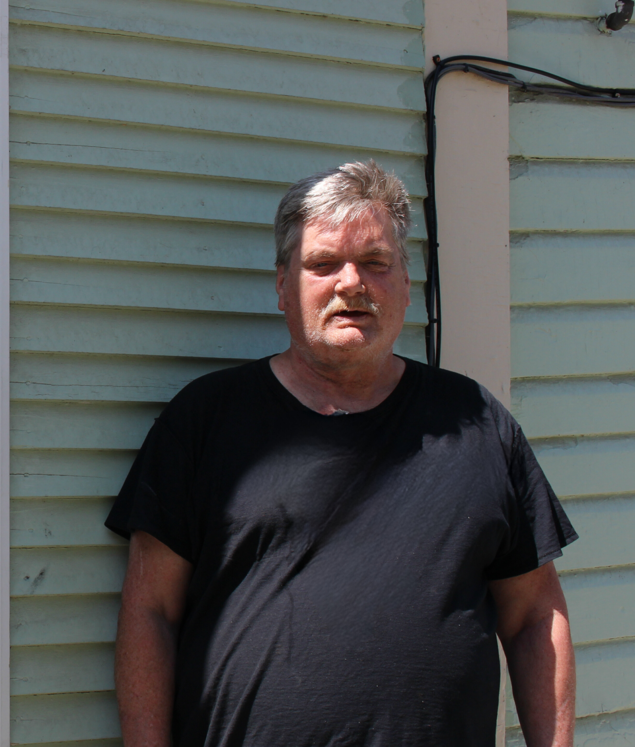 A man standing outside with a black tee shirt and short hair. He is standing in front of a wall that is a light teal color.