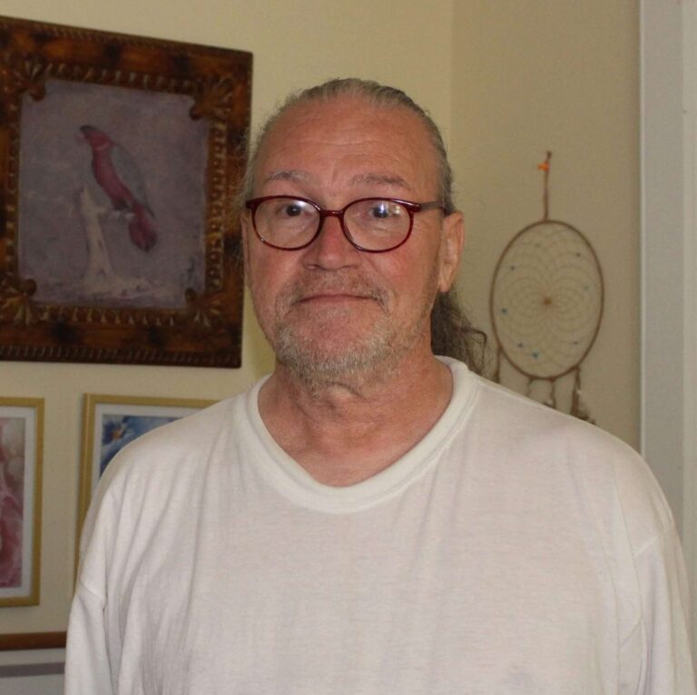 A man wearing glasses, a white tee shirt, and a low pony tail is standing in a room with artwork on the walls behind him