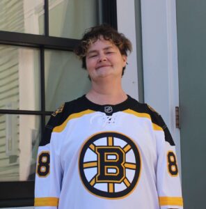 Photo of someone wearing a Boston Bruins jersey. They have short brown hair and are smiling, standing outside in front of windows.