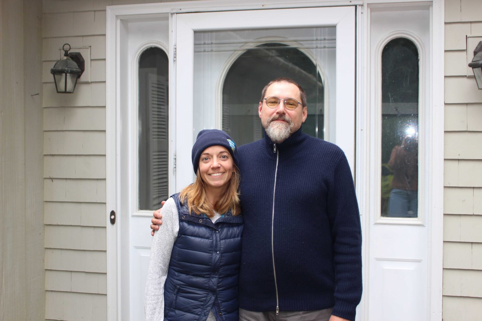 Photo of a man and woman standing in front of a white door to a home. The woman is wearing a blue beanie and vest. The man is wearing a dark sweater.