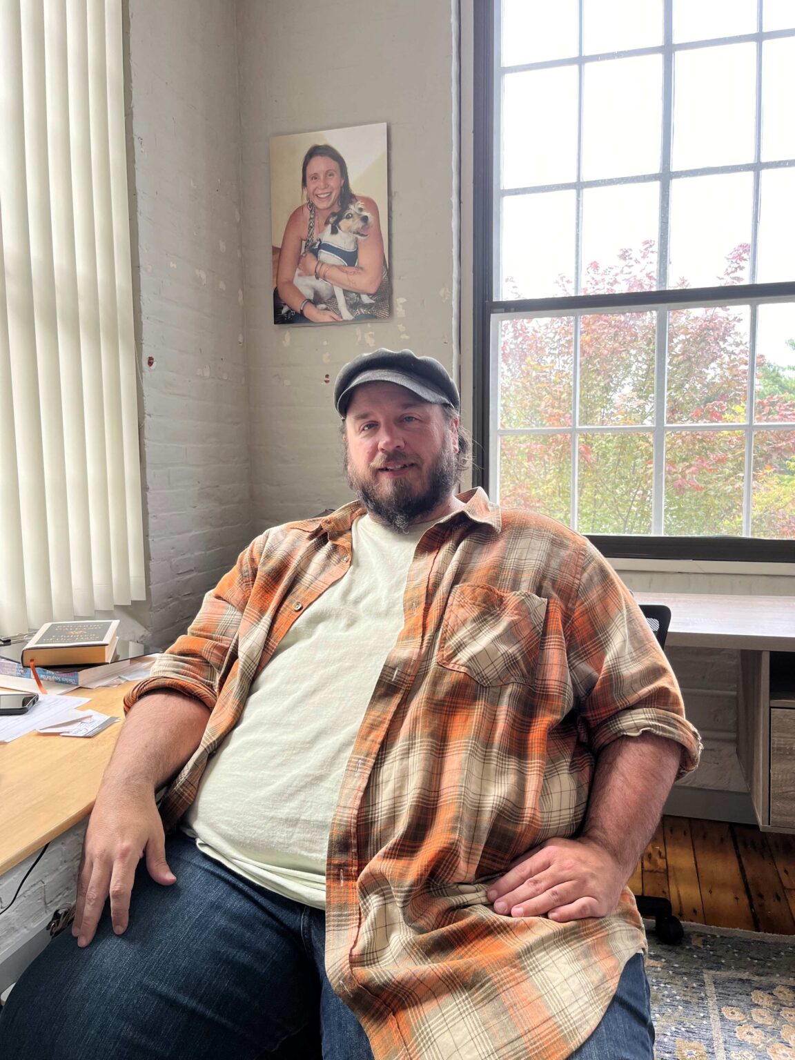 Photo of a man sitting on an office chair smiling. He is wearing an orange and red flannel and a hat.
