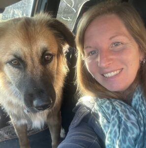 A selfie of a woman and her dog. They are both sitting in a car. The woman in the photo is wearing a blue shirt and cheetah patterned blue scarf. The dog has brown fur