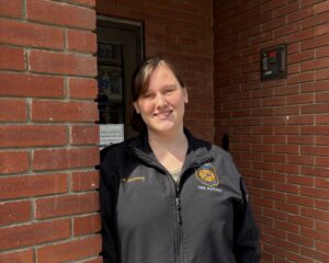 A woman stands in a brick door way. She is wearing a black shirt and has her hair in a pony tail.