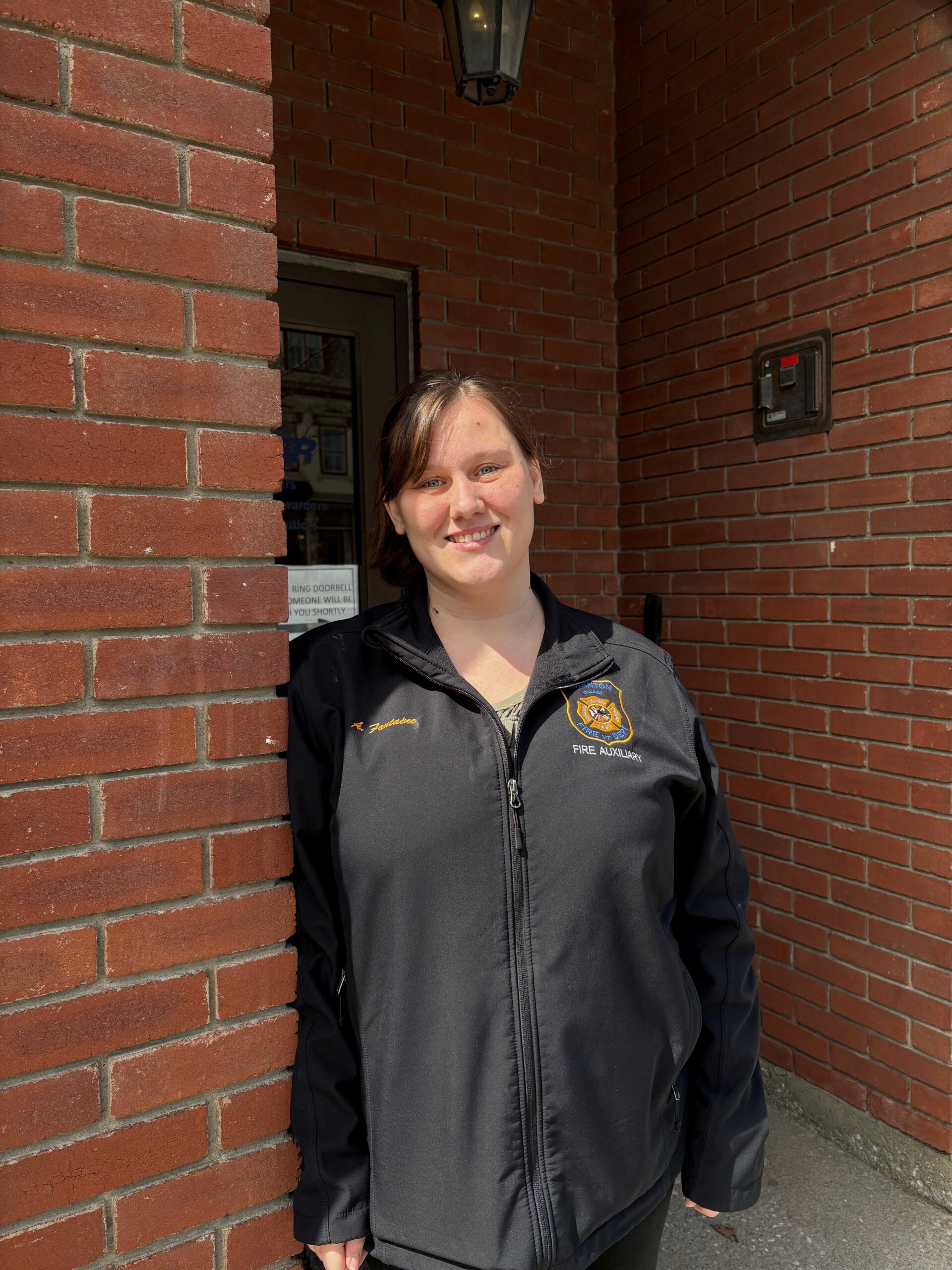 A woman stands in a brick door way. She is wearing a black shirt and has her hair in a pony tail.